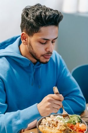 Bearded young ethnic male in blue hoodie tasting fresh delicious food in plate on blurred background